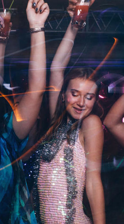 Group of women dancing and raising cocktails on a nightclub dance floor with sequined outfits and colorful party lights