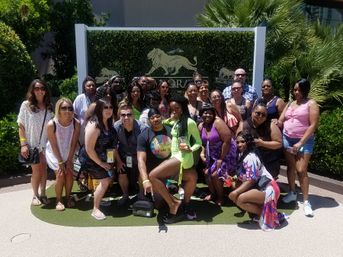 Smiling group of people in summer outfits posing for a sunny resort group photo in front of a decorative lion-emblem sign and tropical landscaping
