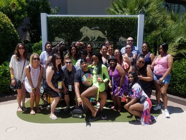 Smiling group of people in summer outfits posing for a sunny resort group photo in front of a decorative lion-emblem sign and tropical landscaping