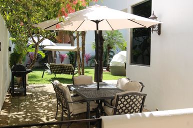 Sunlit backyard patio with a metal outdoor dining table and cushioned chairs under a large beige umbrella, a charcoal grill on pavers, green lawn with lounge chairs and tropical plants in a private garden courtyard.