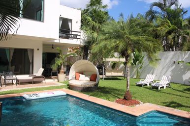 Sunny tropical villa backyard with turquoise swimming pool, round canopy daybed with orange pillows, palm trees, green lawn and striped lounge chairs.