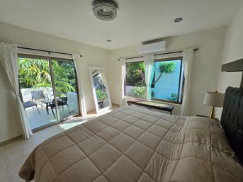 Sunlit modern bedroom with a beige quilted comforter, sliding glass doors opening to a patio with chairs, large window with white curtains, full-length mirror, bench, wall-mounted AC, and lush tropical greenery outside.