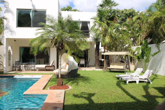 Tropical backyard with turquoise swimming pool and terracotta edge, modern white two-story house, palm trees, cushioned round daybed, swing bench and striped lounge chairs on a green lawn.