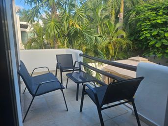Tropical balcony with four modern black mesh patio chairs and a glass-top side table on tiled floor, white stucco railing overlooking lush palm trees and dense green foliage — sunny outdoor seating area.
