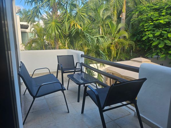 Tropical balcony with four modern black mesh patio chairs and a glass-top side table on tiled floor, white stucco railing overlooking lush palm trees and dense green foliage — sunny outdoor seating area.