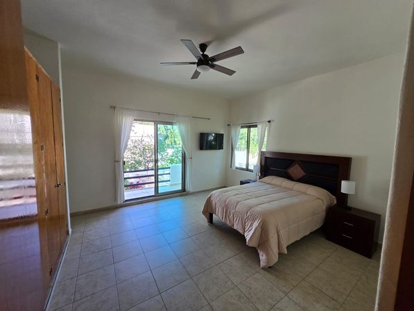 Airy, sunlit bedroom with tiled floor, queen bed with dark wood headboard and matching nightstands, ceiling fan, wall-mounted TV, and sliding glass door with sheer curtains opening to a leafy balcony.