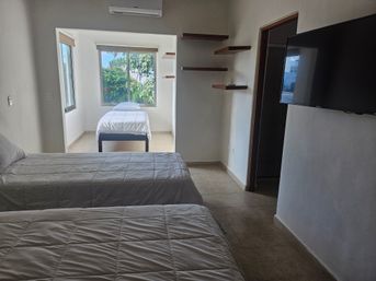 Bright, airy hotel room with two twin beds in the foreground and a single bed in a sunlit alcove by large windows overlooking lush greenery, wall-mounted TV, floating shelves, tile floor and air conditioner.