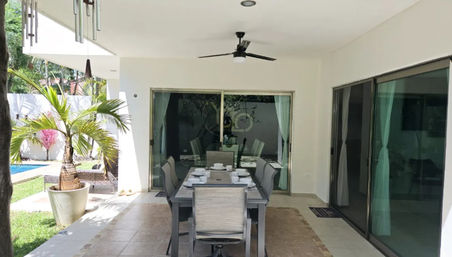 Modern covered patio with six-seat dining table, ceiling fan and sliding glass doors opening to a tropical poolside backyard with potted palm