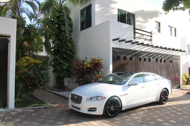 White luxury sedan parked in a sunlit driveway beside a modern white house with a dark wooden garage door, palm trees and tropical landscaping.