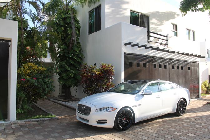 White luxury sedan parked in a sunlit driveway beside a modern white house with a dark wooden garage door, palm trees and tropical landscaping.