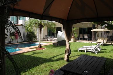 View from a shaded gazebo over a tropical backyard courtyard with a turquoise swimming pool, palm trees, green lawn, white loungers, patio dining set and a hanging swing seat.