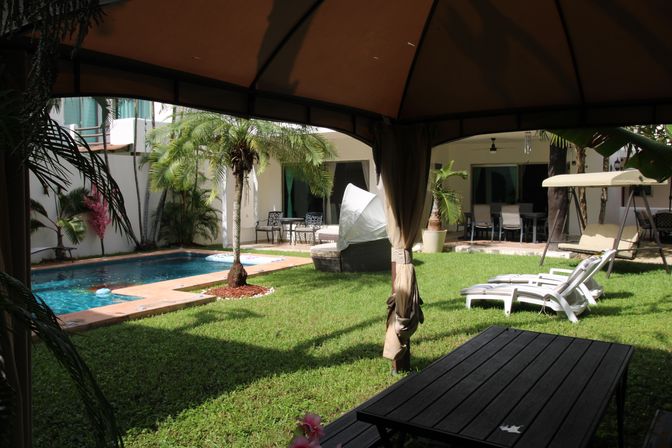 View from a shaded gazebo over a tropical backyard courtyard with a turquoise swimming pool, palm trees, green lawn, white loungers, patio dining set and a hanging swing seat.