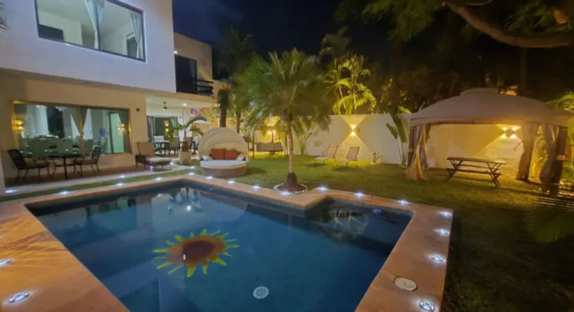 Nighttime tropical backyard with illuminated rectangular pool featuring a sun tile, palm trees, a round daybed, cabana with picnic table, lounge chairs and modern two-story villa with outdoor seating.