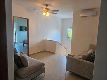 Bright upstairs living area with neutral gray sofas, tiled floor, ceiling fan and split AC, small window with palm view and open doorway to bedroom with wall-mounted TV