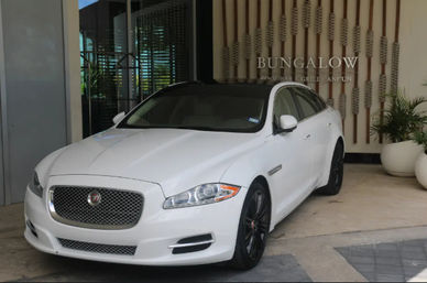 Sleek white Jaguar luxury sedan parked at a resort entrance next to decorative wooden wall panels and large potted plants, black wheels and panoramic roof visible.