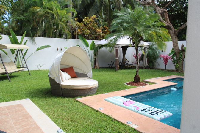 Tropical backyard pool with turquoise water, inflatable lounger, round wicker canopy daybed with cushions on a manicured lawn, palm trees and a shaded cabana by a white privacy wall.