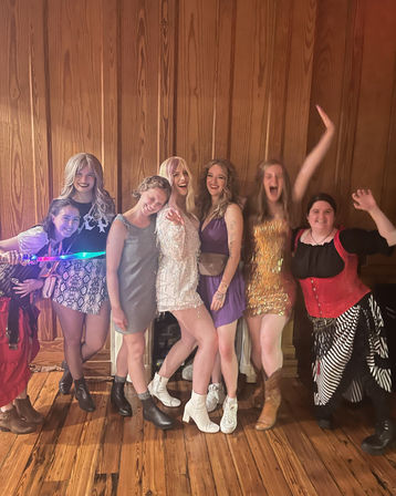 Seven friends in colorful party outfits and sparkly dresses posing and laughing against a wooden-paneled wall, one holding a glowing toy sword.