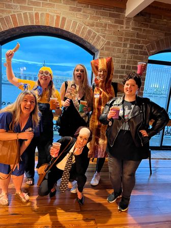 Six people in playful costumes (minion, bacon strip, rocker, and a suit-and-wig gag) holding drinks and posing inside a brick-walled waterfront venue with arched windows at dusk