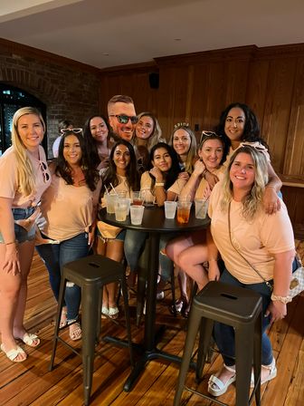 Smiling group of women in matching peach shirts gathered around a high-top table with cocktails in a cozy wood-paneled bar celebrating a bachelorette party.