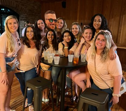 Smiling group of women in matching peach shirts gathered around a high-top table with cocktails in a cozy wood-paneled bar celebrating a bachelorette party.