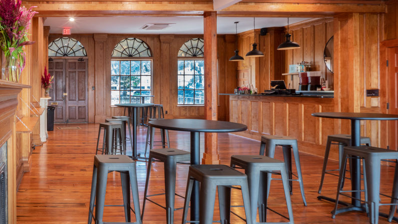 Inviting wood-paneled event lounge with polished hardwood floors, round high-top tables, metal stools, pendant lights and arched windows with a tree-lined view.