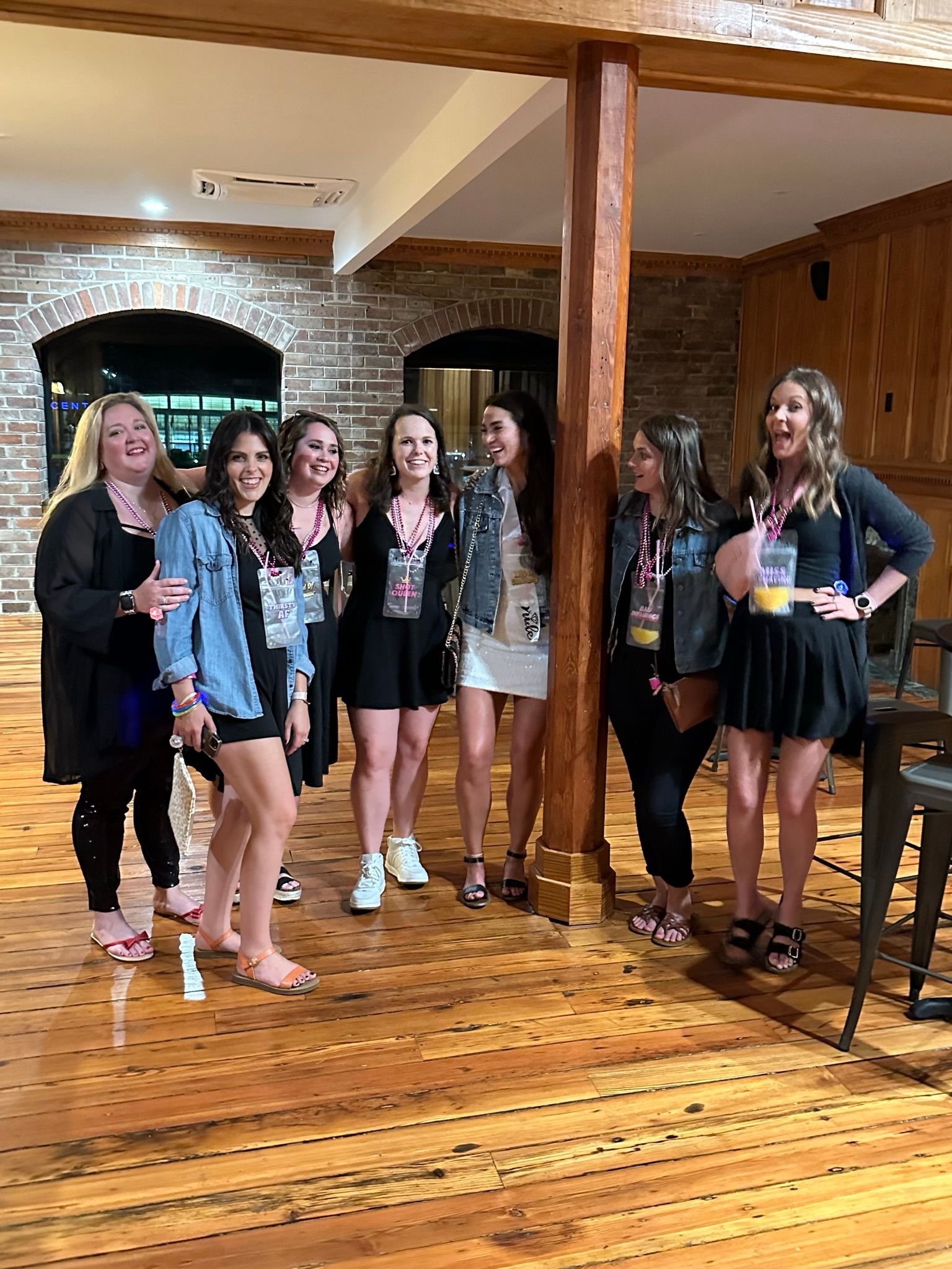 Seven women laughing and posing at a bachelorette party in a rustic indoor venue with exposed brick walls, a wooden support beam and polished hardwood floors, many wearing black dresses, denim jackets, beaded necklaces and a sash.