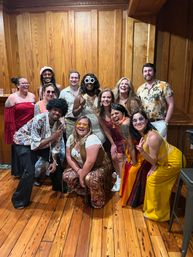 Thirteen friends in colorful 1970s‑inspired outfits and sunglasses posing and laughing on a polished hardwood floor in a warm wood‑paneled room — retro costume party group photo.