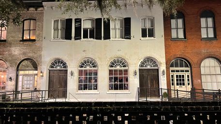 Charming nighttime street view of historic brick and white-painted row houses with arched windows, wooden doors, black shutters, lantern lights, and Spanish moss-draped trees