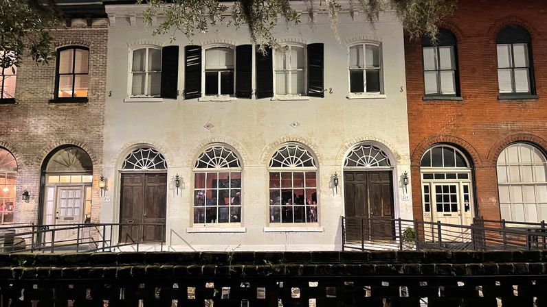 Charming nighttime street view of historic brick and white-painted row houses with arched windows, wooden doors, black shutters, lantern lights, and Spanish moss-draped trees