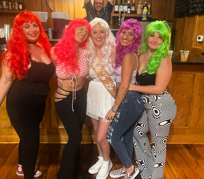 Five women wearing colorful wigs (red, pink, white, purple, green) and a bride-to-be sash pose inside a wood-paneled bar for a playful night out, with a man photobombing in the mirror behind them.
