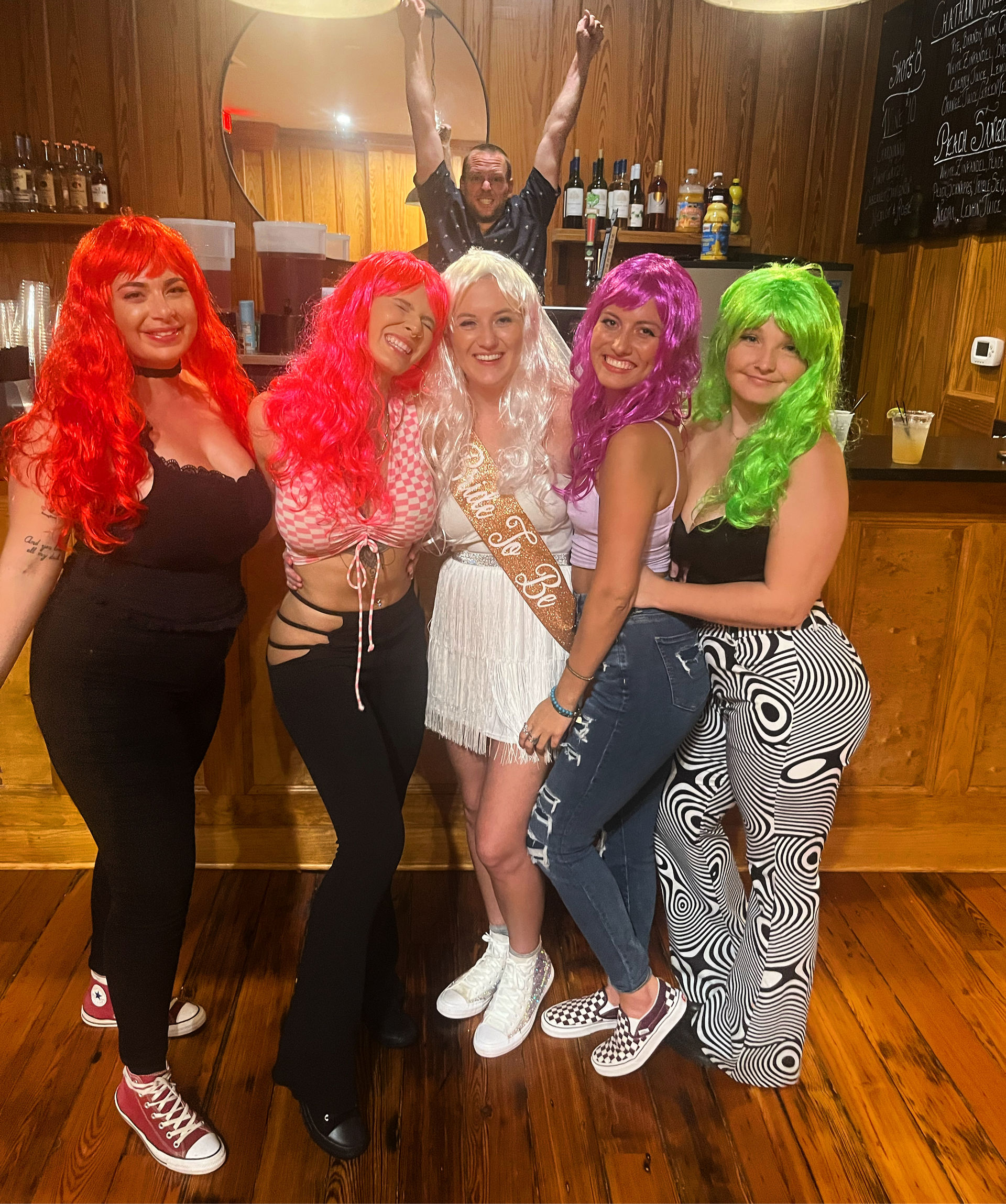 Five women wearing colorful wigs (red, pink, white, purple, green) and a bride-to-be sash pose inside a wood-paneled bar for a playful night out, with a man photobombing in the mirror behind them.
