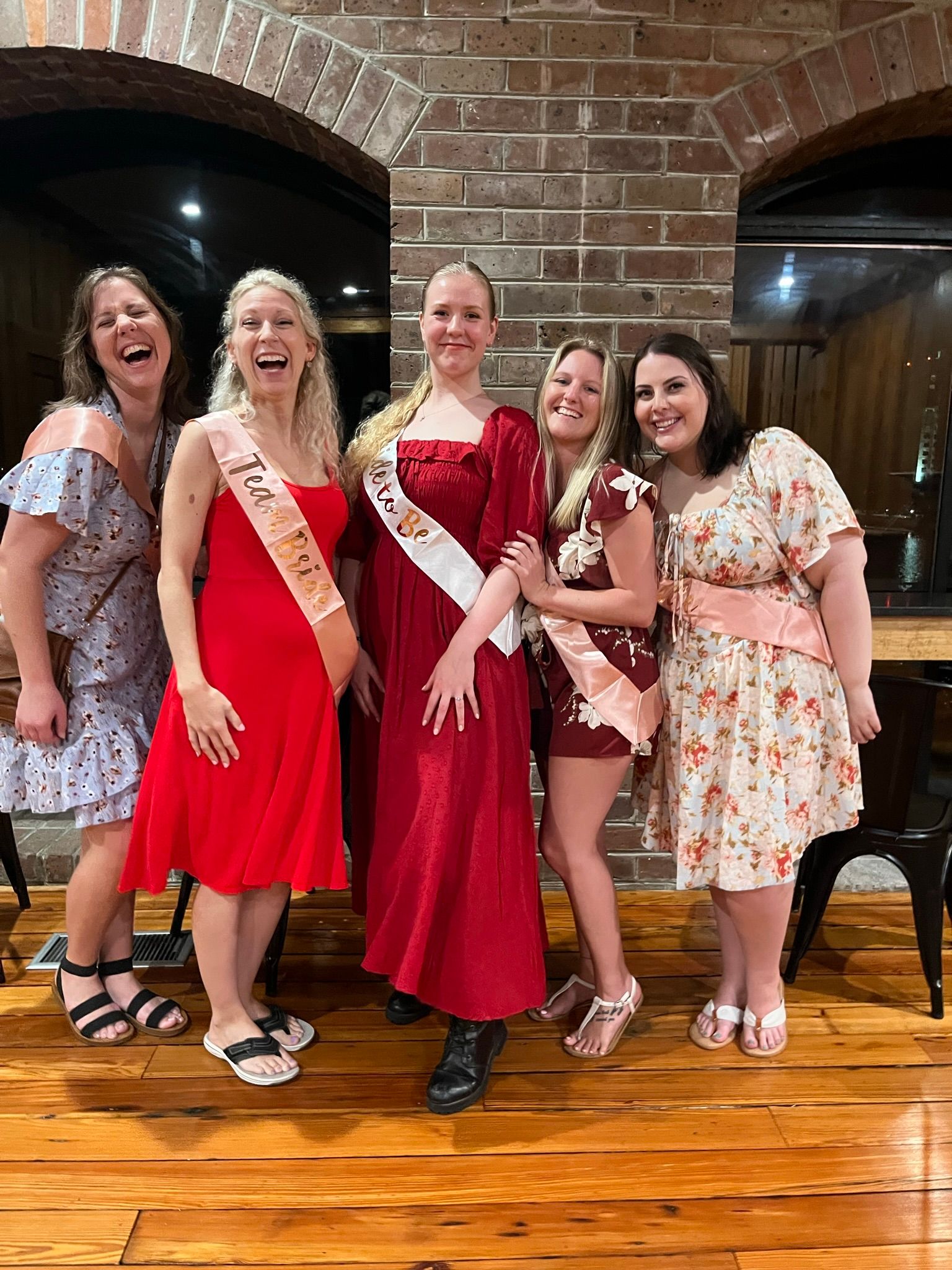Five friends in dresses and pink sashes laughing and posing at a bachelorette party in a rustic brick-walled indoor venue with polished wood floors.