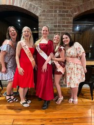 Five friends in dresses and pink sashes laughing and posing at a bachelorette party in a rustic brick-walled indoor venue with polished wood floors.