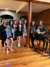 Seven friends posing indoors at a rustic venue with hardwood floors and exposed brick, dressed for a bachelorette/girls' night out with bead necklaces and lanyards.