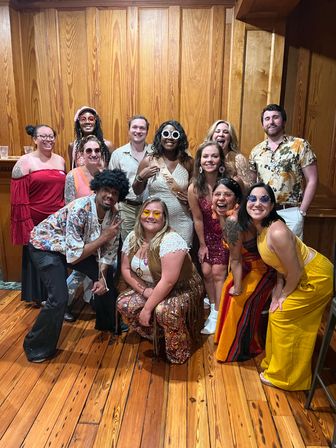 Smiling group of friends in colorful 1970s-inspired outfits posing for a bachelorette party photo in a wood-paneled room with hardwood floor