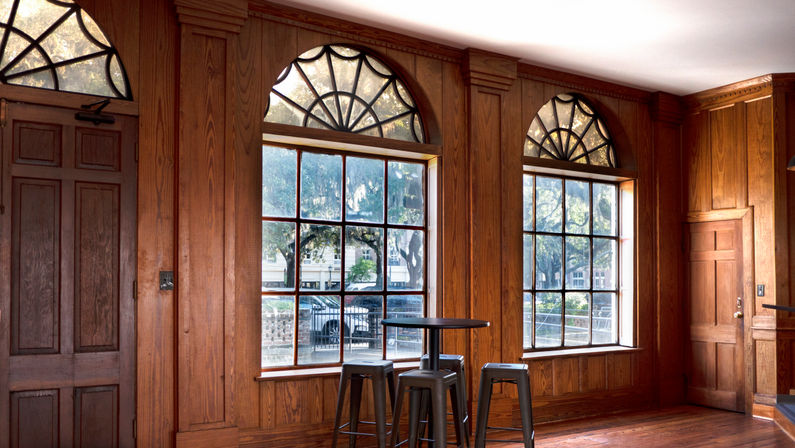 Cozy sunlit wood-paneled room with three arched transom windows and multi-pane glass overlooking a tree-lined street, furnished with a round high table and metal bar stools.