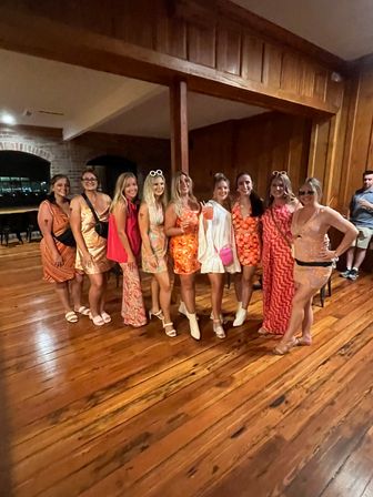 Group of women in bright orange and pink dresses and sundresses posing and smiling in a wood‑paneled hall with polished hardwood floors, some wearing sunglasses or white boots and holding drinks — lively indoor party scene.