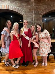 Five smiling women at a bachelorette celebration in a rustic brick-walled venue — bride-to-be in a long red gown with a white sash, friends in red, floral and patterned dresses wearing pink sashes.