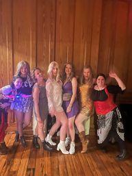 Seven friends in colorful, sparkly party outfits and boots posing and smiling on a wooden floor in front of a wood-paneled wall at an indoor celebration.