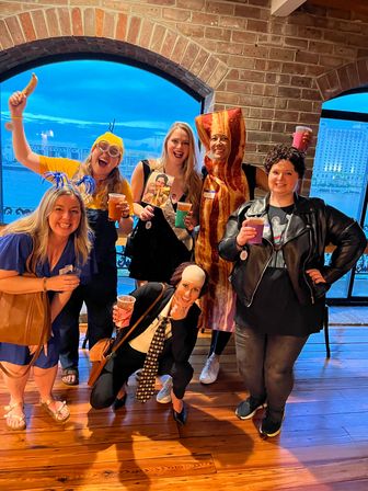 Group of six friends in playful costumes (bacon suit, minion with banana, retro rocker, sash) holding drinks inside a brick-walled, arched-window waterfront venue.