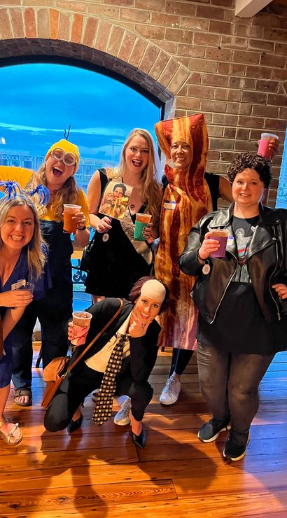 Group of six friends in playful costumes (bacon suit, minion with banana, retro rocker, sash) holding drinks inside a brick-walled, arched-window waterfront venue.