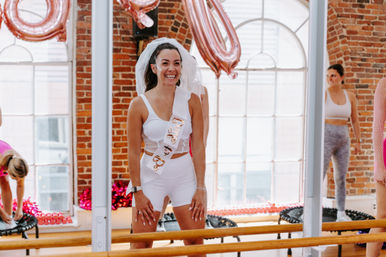 Smiling woman wearing a bridal veil and "bride-to-be" sash participating in a bachelorette rebounder workout in a bright brick-walled studio with arched windows and rose-gold balloons