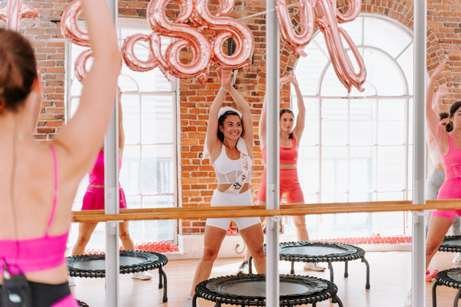 Smiling women in colorful activewear leading a rebounder trampoline fitness class in a mirrored brick studio with rose-gold balloons