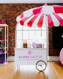 Pink-and-white striped umbrella shading a refreshment cart in an exposed-brick urban loft studio, wooden floors, arched window, champagne bottles and a playful unicorn decoration.