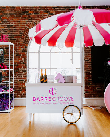 Pink-and-white striped umbrella shading a refreshment cart in an exposed-brick urban loft studio, wooden floors, arched window, champagne bottles and a playful unicorn decoration.