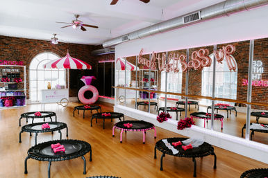 Loft-style urban fitness studio with exposed brick and arched windows, mirrored wall and hardwood floor, mini trampolines set up with pink dumbbells and pom-poms, festive pink balloons and striped canopy for a playful rebound workout.