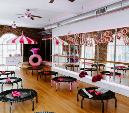 Loft-style urban fitness studio with exposed brick and arched windows, mirrored wall and hardwood floor, mini trampolines set up with pink dumbbells and pom-poms, festive pink balloons and striped canopy for a playful rebound workout.