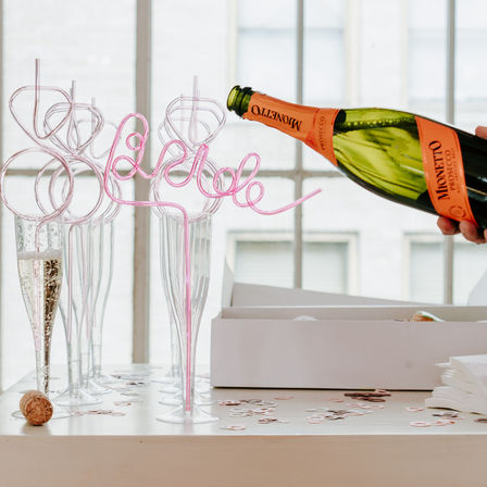 Sparkling wine poured into tall flute glasses with pink curly novelty straws and confetti on a sunlit table by a window