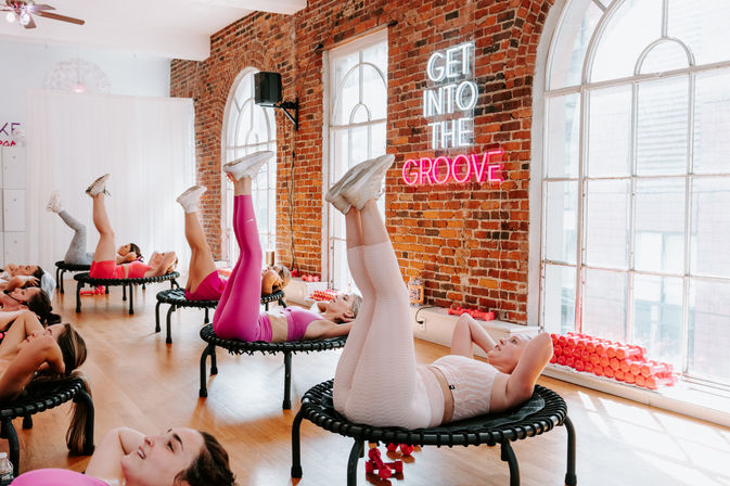 Group rebounder trampoline fitness class in a sunlit brick-walled studio, participants lying on mini trampolines with legs raised, neon “Get Into The Groove” sign, arched windows and stacked pink dumbbells.