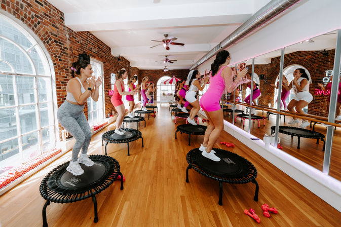 Energetic group rebound class in a bright urban loft fitness studio — participants on mini trampolines wearing pink and white activewear, wood floors, exposed brick, large arched windows and a mirrored wall.